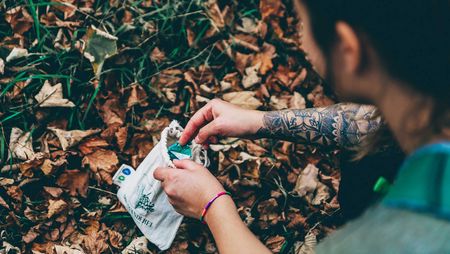 Eine Person sammelt im Wald Müll und steckt diesen in einem Leinensack.