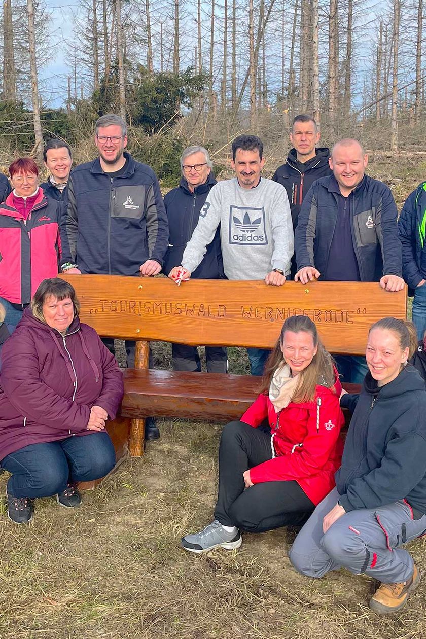 Mitarbeitende der Wernigerode Tourismus GmbH sitzen vor einer Bank am neu gepflanzten Tourismuswald im Stadtforst Wernigerode.